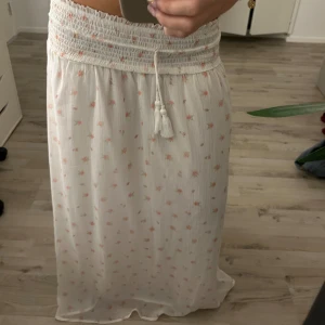 Vit långkjol med små blommor - Superfin vit långkjol med små rosa blommor och bred smock i midjan. Kjolen har knytband med tofsar och är gjord i ett lätt, luftigt tyg som känns perfekt för varma dagar. Den har en flowy passform och ger en romantisk vibe.Bortklipt lapp, mellan storlek 158-170.