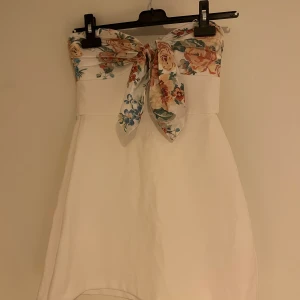 Vit klänning från zara - Superfin vit kortklänning med axelbandslös design och jättefint knyte framtill. Överdelen har blommigt mönster och klänningen har en lätt utsvängd form. Perfekt för sommarens alla tillfällen! 