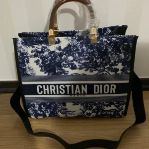 Blåvit axelväska - Säljer en snygg axelväska  i blått och vitt med ikoniskt tryck och tydlig logga framtill. Väskan har både handtag i brunt och axelrem, samt guldfärgade metalldetaljer och dragkedja. Perfekt för dig som vill sticka ut med en lyxig och trendig väska. Helt ny. 