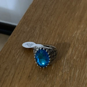 Statementring med blå sten, storlek 20 - Cool silverfärgad ring med en stor, ovalslipad blå sten i mitten. Ringen har detaljerade mönster längs bandet och en glittrig känsla tack vare den facetterade stenen. Perfekt för dig som vill sticka ut med accessoarer.