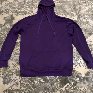 Lila hoodie med huva och magficka - Säljer en stilren lila hoodie med huva och dragsnören. Tröjan har en klassisk magficka och ribbade muddar vid ärmslut och nederkant. Perfekt för dig som gillar en enkel och snygg look.