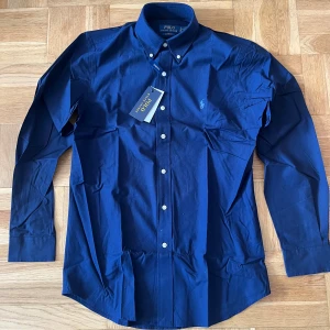 Marinblå skjorta från Polo Ralph Lauren - Snygg marinblå skjorta från Polo Ralph Lauren i custom fit med stretch. Skjortan har klassisk button down-krage, vita knappar och broderad logga på bröstet. Tillverkad i bomull med stretch för extra komfort. Perfekt för dig som gillar stilrena och tidlösa plagg.