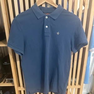 Blå pikétröja från Morris - Snygg blå pikétröja från Morris med klassisk krage, tre knappar och broderad logga på bröstet. Tillverkad i bomull för en skön och luftig känsla. Perfekt till jeans eller shorts för en chill och stilren look. Storlek Small. 