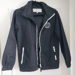 Svart vindjacka med NY patch,barn jacka 12 år - Svart vindjacka från ID Identity med vit dragkedja och två fickor framtill. Jackan har en snygg NY-patch på bröstet och elastiska muddar vid ärmslut och midja. Perfekt för blåsiga dagar och enkel att matcha med streetwear.