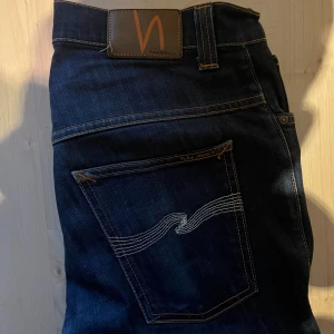Mörkblå jeans från Nudie Jeans Co - Säljer ett par mörkblå jeans från Nudie Jeans Co i klassisk rak modell. Jeansen har snygga kontrastsömmar, ikonisk våg på bakfickan och är tillverkade i slitstark bomull. Perfekta för dig som gillar stilrena och tidlösa jeans.