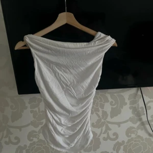 Vit offshoulder kortklänning - Snygg vit kortklänning med offshoulder-design och draperade detaljer längs sidorna. Klänningen har en figurnära passform och är tillverkad i ett mjukt, stretchigt material som ger en cool och modern look. Perfekt för dig som gillar stilrena och trendiga plagg.