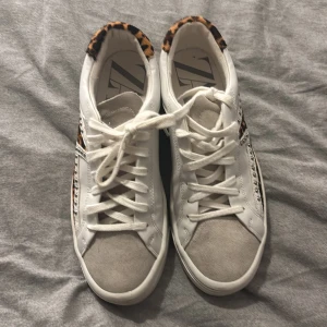 Leopardmönstrade sneakers med nitar - Snygga vita sneakers från Zara i storlek 39 med leopardmönstrade detaljer på hälen och sidan. Skorna har silverfärgade nitar längs sidorna, snörning och en rund tå i beige mocka. Perfekt för dig som gillar att sticka ut med coola detaljer och djurmönster.