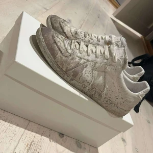 Äkta Maison margiela skor  - Säljer ett par unika maison margiela sneakers i vit och beige canvas med distressed och sliten look. Skorna har snörning, rund tå och platt sula. Råa kanter och vintage-vibbar gör dem till ett statement-piece för dig som gillar edgy mode.dom har lite hål i skon så ni vet men inget man tänker på 