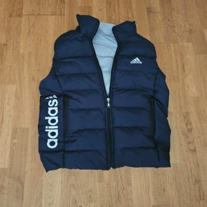 Svart dunväst från Adidas - Snygg svart dunväst från Adidas i storlek M. Västen har en quiltad design, dragkedja framtill och två fickor med dragkedja. Adidas-logga på bröstet och stor vit logga längs sidan ger en sportig vibe. Perfekt för lager-på-lager-stil under kyliga dagar. Använt bara några gånger och pris kan diskuteras.