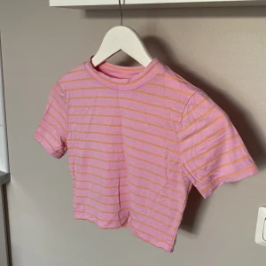 Rosa croppad t-shirt från Collusion - Säljer en croppad t-shirt från Collusion i rosa med tunna orangea ränder, storlek 36. T-shirten har korta ärmar och rund halsringning. Materialet är mjukt och stretchigt med inslag av polyester.