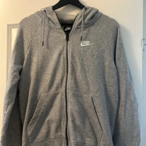 Grå Nike hoodie med dragkedja - Klassisk grå hoodie från Nike med vit logga på bröstet. Tröjan har huva med snörning, hel dragkedja och två stora fickor framtill. Perfekt för chill dagar eller när du vill ha en sportig look. Mjuk och skön i bomullsmix.