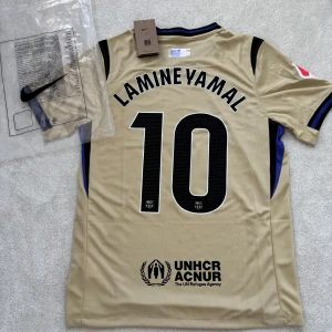 FC Barcelona Lamine Yamal x kobe bryant - FC Barcelona fotbollströja i beige med svarta detaljer, Lamine Yamal och nummer 10 på ryggen. Nike-logga, klubbmärke och sponsortryck på bröstet. Kortärmad modell i lätt och ventilerande funktionsmaterial. Snygga blå och svarta ränder på ärmslut och krage.