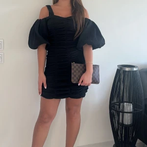 Svart kort klänning med puffärmar - Supersnygg svart kort klänning med markerade puffärmar och offshoulder-design. Klänningen har rynkade detaljer längs hela kroppen och en figurnära passform som framhäver siluetten. Perfekt för dig som vill sticka ut med en trendig och elegant look.