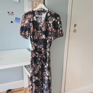 Blommig långklänning från KappAhl - Supersnygg långklänning från KappAhl med svart botten och blommigt mönster i beige, rosa och vitt. Klänningen har korta puffärmar, knappar framtill, knytskärp i midjan och diskreta paljettdetaljer som ger extra glans. Perfekt för dig som vill sticka ut lite extra.