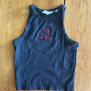 Mörkgrå Hello Kitty ribbad linnetopp - Säljer en mörkblå ribbad linnetopp från H&M med röd Hello Kitty-brodyr framtill. Toppen är ärmlös och har en tight passform med hög halsringning. Perfekt för dig som gillar söta detaljer och vill sticka ut med en ikonisk look.