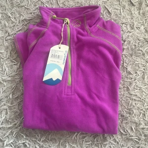 Lila fleece-tröja från Stormberg - Supermjuk lila fleece-tröja från Stormberg med limegröna detaljer och half zip framtill. Perfekt för kyliga dagar och outdoor vibes. Långärmad och med hög krage som håller dig varm. Snygg och sportig look som funkar till allt.