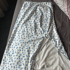 Vit blommig långkjol från SHEIN - Superfin långkjol från SHEIN i vitt med små blå blommor över hela tyget. Kjolen har en snygg slits framtill med snörning som detalj. Skön passform. Perfekt för en söt och somrig look!