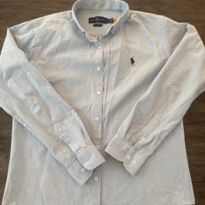 Ljusblå skjorta från Ralph Lauren - Klassisk ljusblå skjorta från Ralph Lauren med broderad logga på bröstet. Skjortan har lång ärm, knappar framtill och krage. Tillverkad i mjuk bomull som känns skön mot huden. Perfekt för en clean och stilren look. Väldigt liten i storleken så passar M och S