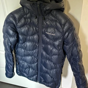 Mörkblå dunjacka Peak Performance - Mycket bra skick! Lite defekt i krage. Mörkblå dunjacka från Peak Performance med vågformat quiltat mönster. Jackan har huva, dragkedja framtill och två sidofickor med dragkedja. Lätt att bära, klassisk logga framtill och på ena ärmen. 