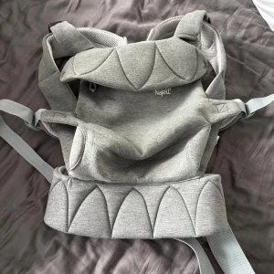 Grå Najell Easy bärsele - Säljer en Najell Easy bärsele i snyggt grått tyg med quiltade detaljer. Selen har breda, vadderade axelband och smarta spännen med mintgröna knappar. Materialet är mjukt och andas, perfekt för att bära små barn på ett bekvämt sätt. Denna är rekomenderad från 3 månader upp till 3 år. Aldrig använd utan endast testad men min dotter tycker inte om att sitta i den därav säljs den. Kan gå ner i pris vid snabb affär.  