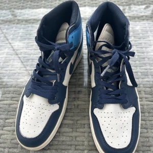 Nike Air Jordan 1 Retro High Obsidian  - Säljer ett par Nike Air Jordan 1 High i blått och vitt skinn med mörkblå detaljer och snörning. Klassisk siluett med perforerad tå och ikonisk Jordan-logga på sidan. Snygg kontrast mellan ljusblå och marinblå paneler, perfekt för dig som gillar streetstyle. Som ny typ