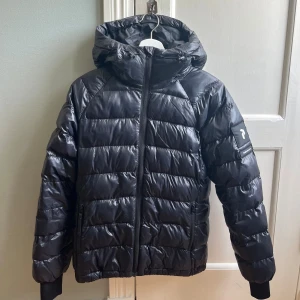 Svart dunjacka från Peak Performance - Snygg svart dunjacka från Peak Performance med huva och dragkedja framtill. Jackan har quiltad design, ribbade muddar och logga på ärmen. Perfekt för kalla dagar och riktigt skön att bära. Klassisk look som funkar till allt.