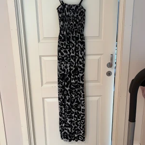 Svartvit leopardmönstrad långklänning  HUAME - Snygg långklänning från HUAME med svartvitt leopardmönster och smockad överdel. Klänningen har smala axelband och ett flowy fall som ger en cool och avslappnad vibe. Perfekt för dig som vill sticka ut med ett djurmönster och ändå hålla det stilrent.