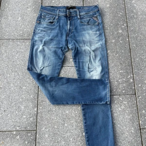 Blå Replay jeans med slitningar - Säljer ett par blå jeans från Replay med snyggt slitna detaljer och en liten reva på vänster lår. Klassisk femficksmodell, normal passform och medelblå tvätt. Perfekta för dig som gillar en avslappnad och trendig look.