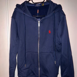 Marinblå hoodie från Polo Ralph Lauren - Snygg marinblå hoodie från Polo Ralph Lauren med dragkedja, huva och snörning. Klassisk röd logga broderad på bröstet och två fickor framtill. Perfekt för en chill och stilren look.