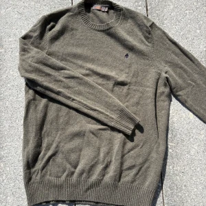 Olivgrön stickad tröja från Morris - Säljer en olivgrön stickad tröja från Morris med rund halsringning och diskret broderad logga på bröstet. Tröjan har ribbade muddar vid ärmslut och nederkant, perfekt för lager-på-lager under kyliga dagar.