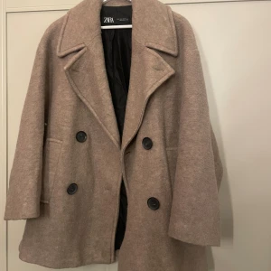 Beige ullkappa från Zara, stl S - Snygg beige kappa från Zara i storlek S. Jackan har dubbelknäppning med stora svarta knappar, bred krage och två fickor framtill. Materialet är en mix av ull och polyester som ger en mjuk och varm känsla. Perfekt för kyliga dagar och en stilren look.