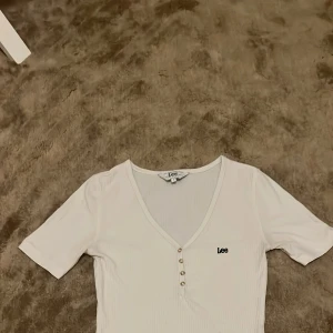 Vit ribbad topp från Lee med knappar - Supersnygg vit ribbad topp från Lee med v-ringning och fyra dekorativa knappar framtill. Kortärmad och figurnära passform som sitter perfekt. Liten Lee-logga broderad på bröstet. Perfekt till jeans eller kjol för en clean look.