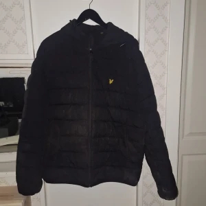 Svart pufferjacka från Lyle & Scott - Pris går att diskutera.    Svart pufferjacka från Lyle & Scott. Jackan har huva, dragkedja framtill och två sidofickor. Den är vadderad och har en klassisk quiltad look. Lite smutsig då vår torktumlare har gått sönder och jag ville inte att dunet i skulle klumpa ihop sig om man tvättade den I tvättmaskinen. 