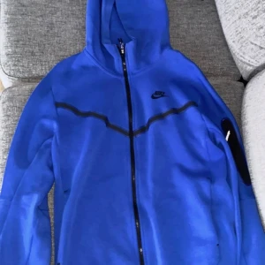 Blå Nike Tech hoodie - Snygg blå Nike Tech hoodie. Den är lite nopprig vid dragkedjan därav lägre pris