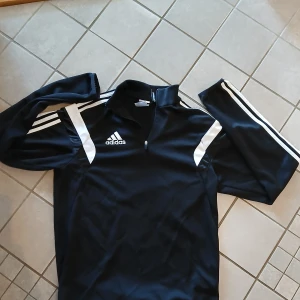 Svart Adidas half zip tröja - Svart långärmad Adidas-tröja med vita ränder på ärmarna och vita detaljer på axlarna. Klassisk Adidas-logga på bröstet och praktisk half zip vid halsen. Perfekt för träning eller sportiga outfits.