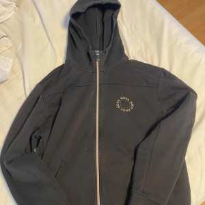 Svart hoodie med dragkedja från BOSS - Säljer en svart hoodie från BOSS med guldfärgat tryck på bröstet. Tröjan har huva, hel dragkedja och två fickor framtill. Tillverkad i mjuk bomullsmix som är skön att ha på sig. 