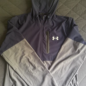 Under Armour vindjacka, mörkblå och grå - Snygg vindjacka från Under Armour i mörkblått/marinblått och grått. Jackan har huva, dragkedja framtill och en bröstficka med dragkedja. Perfekt för dig som gillar sportig stil och vill ha något lätt och funktionellt. Perfekt nu när hösten är påväg också. Kan lösa billigare pris vid snabbaffär.
