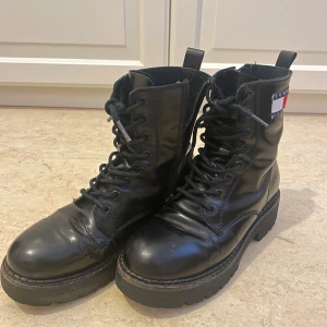 Svarta boots från Tommy Hilfiger - Snygga svarta boots från Tommy Hilfiger med grov sula och snörning framtill. Tillverkade i skinn med rund tå och coolt Tommy Jeans-märke på sidan. Perfekta för dig som vill ha en edgy och urban look.