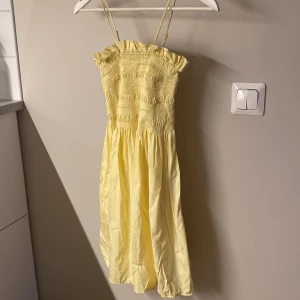 Gul sommarklänning från H&M - Supersöt ljusgul klänning från H&M med smockad överdel, volangkant upptill och tunna justerbara axelband. Tillverkad i 100% bomull och har en luftig, flowy kjol som passar perfekt för varma dagar.