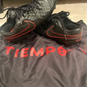 Nike Tiempo svarta fotbollsskor - Svarta Nike Tiempo fotbollsskor med röda detaljer och klassisk Swoosh-logga. Ovandel i skinn med diamantmönstrad struktur och snörning. Yttersula med fasta dobbar för bra grepp på planen. Snygg och sportig look för dig som vill sticka ut på matchen.
