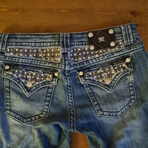 Miss Me jeans med unika detaljer - Säljer ett par blå Miss Me jeans med snygga detaljer i form av bling och broderier på fickorna. Jeansen har raka ben och slitna benslut för en cool look. Klassisk femficksmodell med dekorativa knappar och låg midja.
