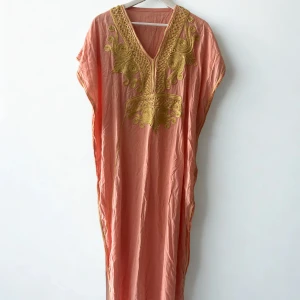 Lång rosa kaftanklänning med broderi - Säljer en lång, luftig kaftanklänning i ljusrosa med vackert guldigt broderi runt v-ringningen och framtill. Klänningen har en loose passform och är ärmlös, perfekt för sommarens varma dagar. Materialet känns lätt och skönt mot huden.