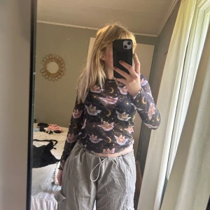 Svart mesh topp med kattmotiv från Monki - Säljer en svart mesh topp från Monki med lång ärm och coolt print av flygande katter, månar och stjärnor i lila och gult. Toppen är genomskinlig och har vågig kant nedtill. Perfekt för dig som gillar unika och lekfulla plagg!