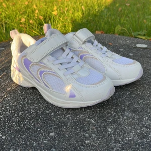 Vita och lila sneakers stl 33 - Snygga vita sneakers med lila detaljer från 7C. Skorna har kardborreband och elastiska snören för enkel påtagning. Ovandelen är i mesh och syntet, vilket ger en sportig look och bra andningsförmåga. Perfekta för dig som gillar stilrena och bekväma skor.