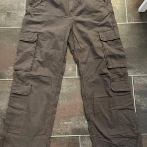 Grå cargobyxor från Bershka - Säljer ett par bruna multicargo-byxor från Bershka i storlek 44. Byxorna har flera stora fickor på benen och klassisk workwear-stil. Tillverkade i bomull med knapp och dragkedja framtill. Perfekta för dig som gillar praktiska och snygga cargopants. Helt oanvända!