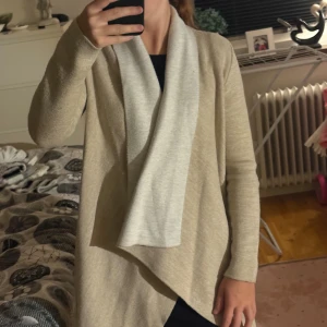 Beige kofta med ljus krage - Säljer en supermjuk och lång beige kofta med öppen front och ljusare krage. Koftan har lång ärm och en avslappnad, loose passform som ger en chill vibe. Perfekt att slänga på sig över en t-shirt eller klänning när det är lite kyligt ute.