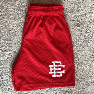 Eric Emanuel shorts  - Säljer ett par röda mesh shorts från Eric Emanuel i storlek medium. De har elastisk midja med vit snörning och ett stort vitt EE-logotryck på vänster ben. Perfekta för sport eller chill, tillverkade i luftig polyester. Shortsen skick är 10-10 knappt använda då dom inte är min still längre, ny pris 2800 på StockX mitt pris 1400 man får också med original påsen. Skriv vid mer frågor.