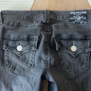True Religion Joey Low Rise Flare Jeans - Säljer ett par mörkgrå True Religion Joey jeans med låg midja och flare/bootcut. Klassiska fickor med lock och silverfärgade knappar bak, samt tydliga sömmar. Riktigt snygga och ikoniska jeans med cool passform.