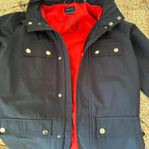 Marinblå  jacka från GANT mm  - Snygg vår/höstjacka från GANT med luva och röd insida och silverfärgade knappar. Jackan har fyra stora fickor framtill och dragkedja. Perfekt för dig som vill ha en stilren och funktionell jacka med streetkänsla.