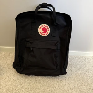 Svart Kånken ryggsäck från Fjällräven - Klassisk svart Kånken ryggsäck från Fjällräven med ikonisk rund röd-vit logga framtill. Ryggsäcken har ett stort huvudfack, framficka med dragkedja och två sidofickor. Tillverkad i slitstark syntet med bärhandtag och justerbara axelremmar. (Väskan är inte äkta men jag ser ingen skillnad)